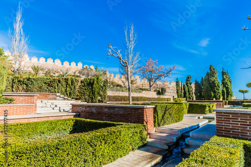 Alcazaba garden with green bushes, some trees, its irrigation canal with the fountain and the stone wall of the fortress in the background, wonderful day with a blue sky in Almería, Spain © Emile