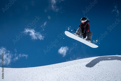 A professional snowboarder girl in flight after jumping from a snow eaves makes a rake against blue sky in sunny day. The concept of winter sports freeride and freestyle