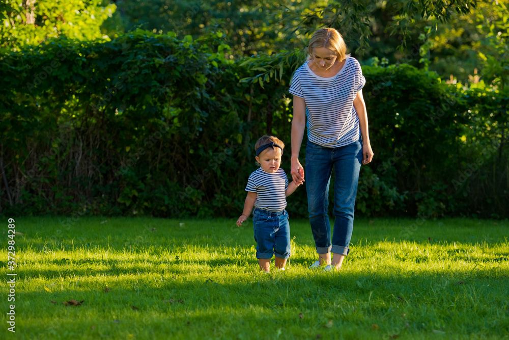 Fototapeta premium A young mother holds the hand of her little daughter on a green lawn in her garden.
