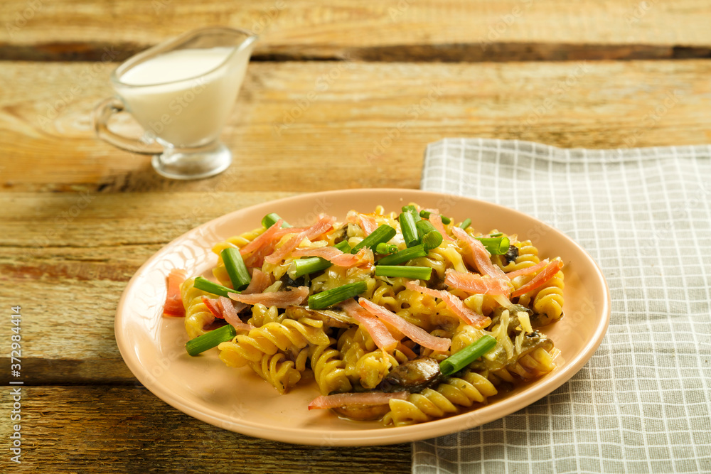 a plate with pasta with ham and mushrooms in a creamy sauce on the table with a fork on a napkin next to the cream. a plate with pasta with ham and mushrooms in a creamy sauce on the table with a fork on a napkin next to the cream.