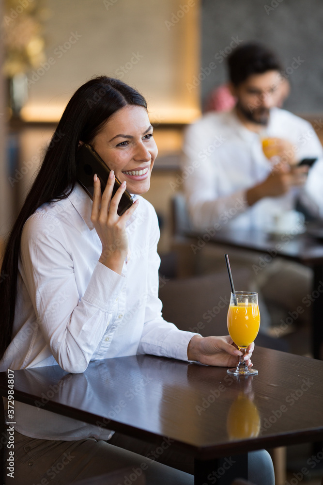 A young woman is sitting at a table in a restaurant, talking on a mobile phone, smiling and drinking orange juice, a young gentleman is sitting behind her and looking at the phone A young woman is sitting at a table in a restaurant, talking on a mobile phone, smiling and drinking orange juice, a young gentleman is sitting behind her and looking at the phone