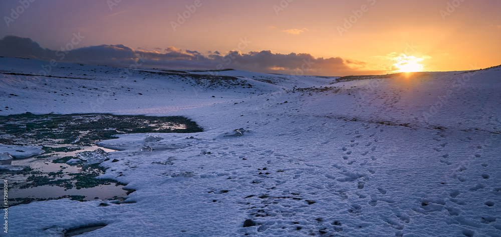 Dramatic sunset in the small mountains covered with snow and foodprints, ice and  water around 