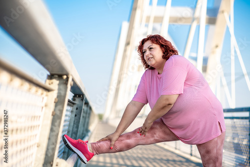 Side image of an obese woman does exercises to warm up her legs on the bridge, preparations for jogging