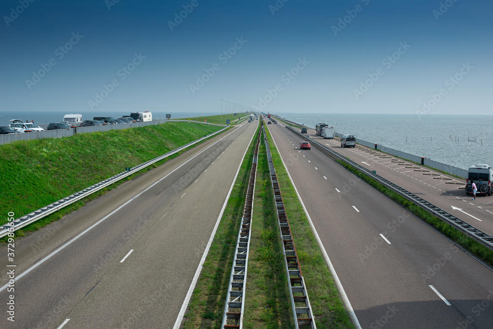 Straight asphalt road. Major dam with causeway,  green grass and cars travelling the road. Sea, blue sky as background.