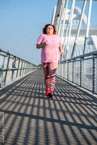 Image of an obese woman jogging on the bridge on a sunny day, doing weight loss training