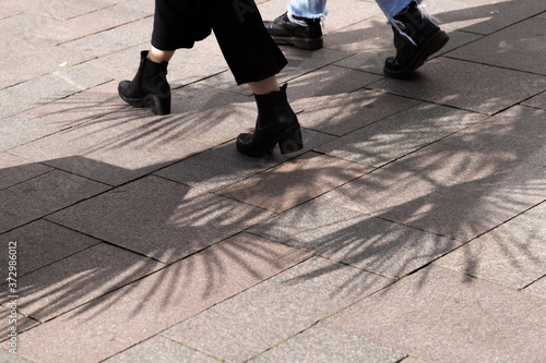 Closeup of the feet of two young females in a shopping district with the shadow pattern of palm trees in the foreground on the pavement
