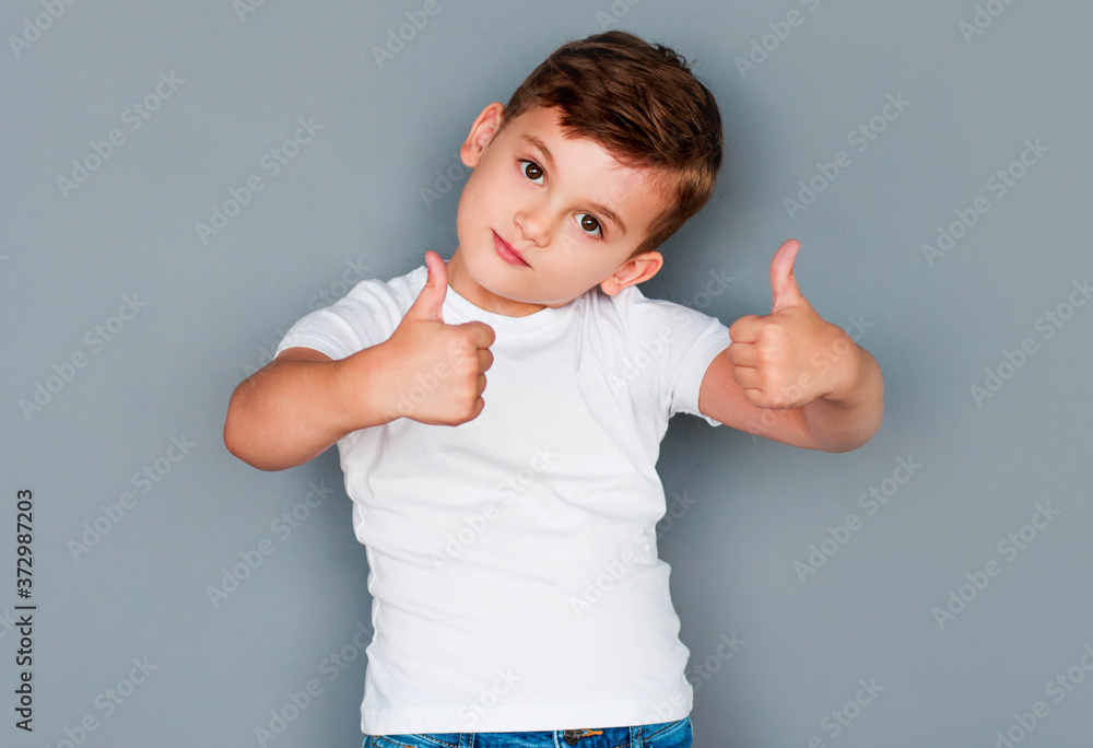 Like gesture. Approval sign. Cheerful boy in white t-shirt showing thumbs up isolated on gray background. Like gesture. Approval sign. Cheerful boy in white t-shirt showing thumbs up isolated on gray background.