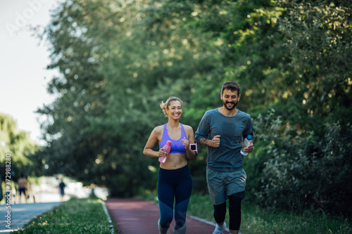 Front image of a young couple jogging on the treadmill on a sunny day, they hold bottles of water in their hands