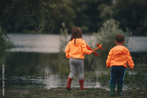 A picture from behind of a brother and sister standing by the river and a girl holding a toy airplane in her hand
