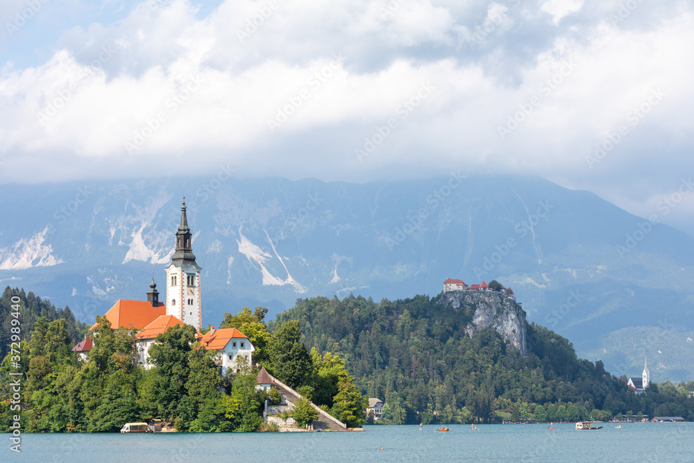 Close up of the iconic Slovenian island on the lake Bled surrounded by trees and vegetation, under a blue summer sky with puffy clouds, with a promontory in the background