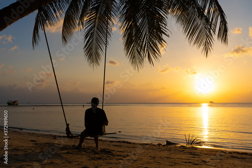 The guy enjoys the sunset riding on a swing on the ptropical beach. Silhouettes of a guy on a swing hanging on a palm tree  watching the sunset in the water.