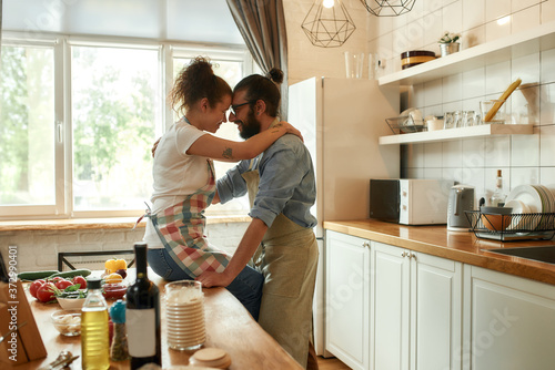 Young man looking at his girlfriend, hugging while standing in the kitchen. Couple embracing while making pizza with vegetables indoors. Cooking together, hobby, lifestyle