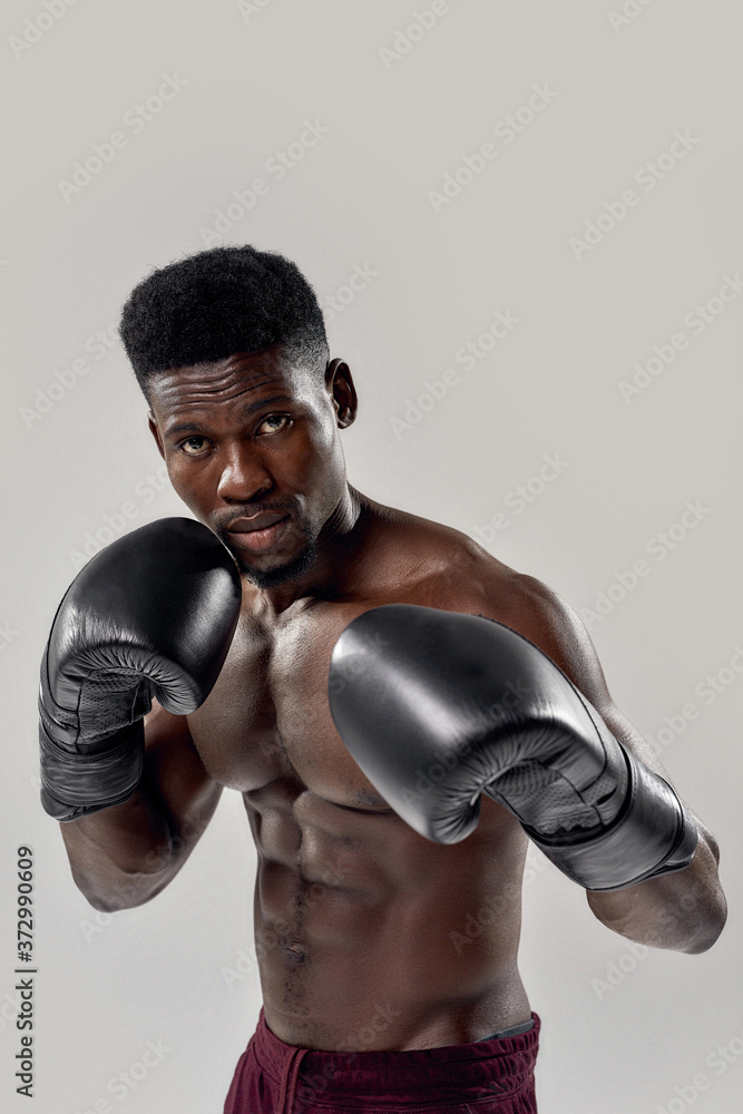 Portrait of young muscular african american male boxer looking at camera, wearing boxing gloves, standing isolated over grey background. Sports, workout, bodybuilding concept
