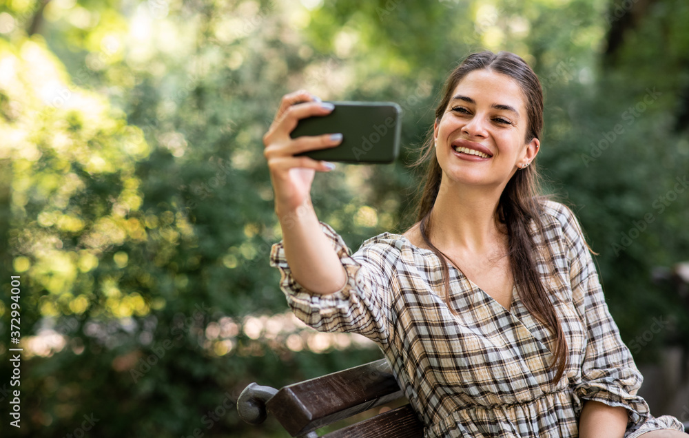 A young woman is sitting on a park bench and taking a picture of herself on her mobile phone on a sunny day