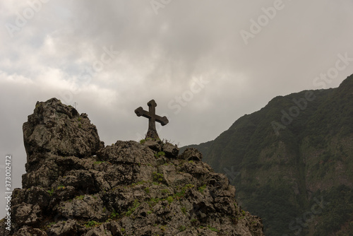Cross on the rock. Typical landscape of Madeira Island. High mountains under cloudy sky. Sao Vicente town. 