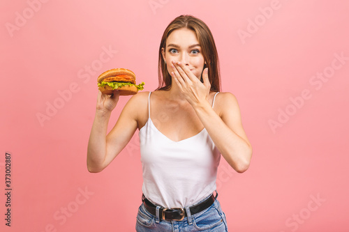Portrait of young beautiful hungry woman eating burger. Isolated portrait of student with fast food over pink background. Diet concept.