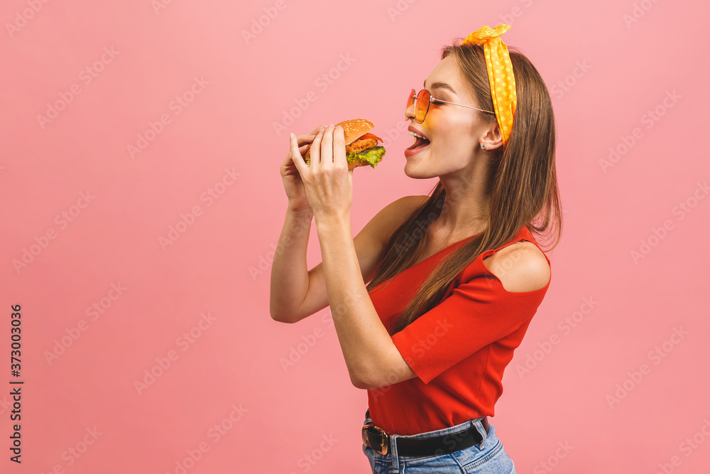 Portrait of young beautiful hungry woman eating burger. Isolated portrait of student with fast food over pink background. Diet concept. Portrait of young beautiful hungry woman eating burger. Isolated portrait of student with fast food over pink background. Diet concept.