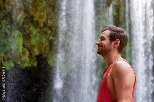 Beautiful shot of a male in a red shirt smiling and standing close to a waterfall photo