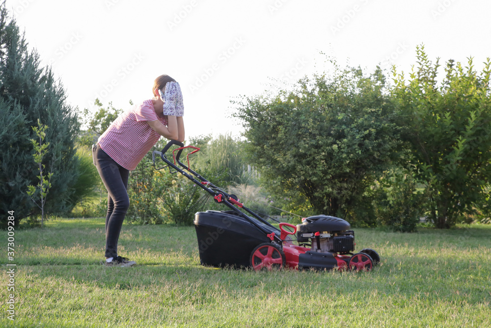 Tired woman making a short break while cutting grass with a lawn mower. Outdoor household chores concept. Tired woman making a short break while cutting grass with a lawn mower. Outdoor household chores concept.