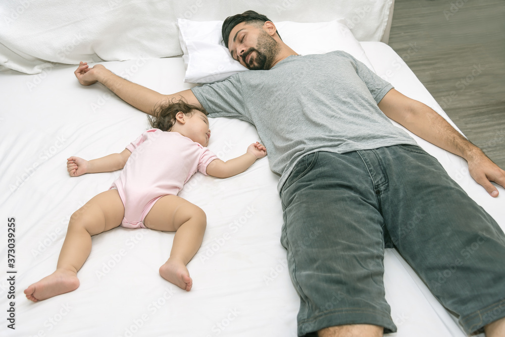 Obraz premium Young handsome father tired sleeping with one year baby girl. Bearded dad sleeps with baby in bed. Little newborn child in white bed. Parenthood ccncept. Love and happiness.