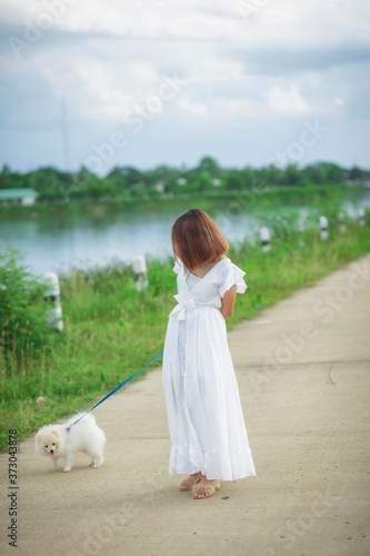 Asian woman in white dress walking with dog in park.