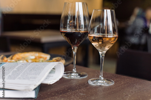 Glasses of red and rose wine, 4 cheese pizza, fresh newspaper on wooden table at the cafe. Dating concept. Weekend couples leisure.