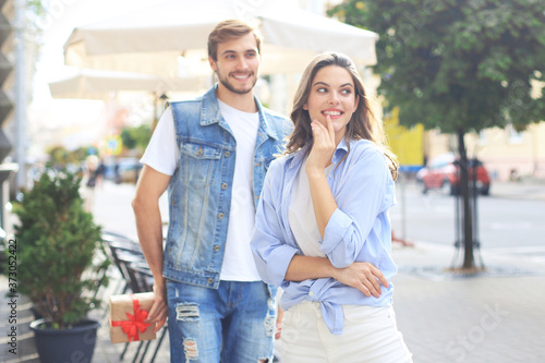 Astonished excited couple in summer clothes smiling and holding present box together while standing on city street. © ty