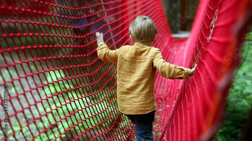 Blonde caucasian boy playing on modern playground nets in the forest. Children activity. Summer traveling male child having fun outdoor. Kids fashion. Healthcare concept. Physical education. Happiness