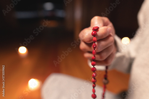 close-up man in clothes for practice and meditation sits in a lotus pose and holds red rosary to concentrate attention in a wooden room with dim light