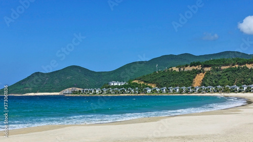 Tropical beach. Curved coastline  turquoise waves with white foam run on the white sand. There are a number of villas and palm trees in the distance. Green mountains against the blue sky. Vietnam.