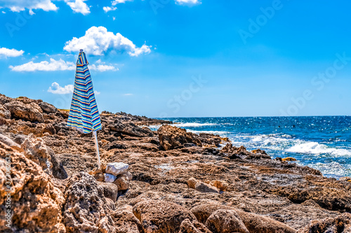 View of a picturesque seascape with rocky cliffs and an umbrella along the Ionian coast of Salento, Puglia region, Italy, Blue sky with clouds and warm colors.  © AlexMastro