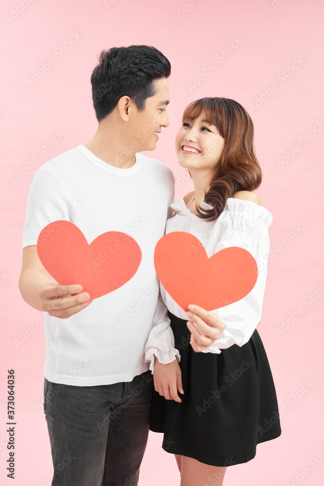 Happy together. Two young cute lovers are looking at the camera and smiling, holding red big paper hearts, embracing on pink background Happy together. Two young cute lovers are looking at the camera and smiling, holding red big paper hearts, embracing on pink background