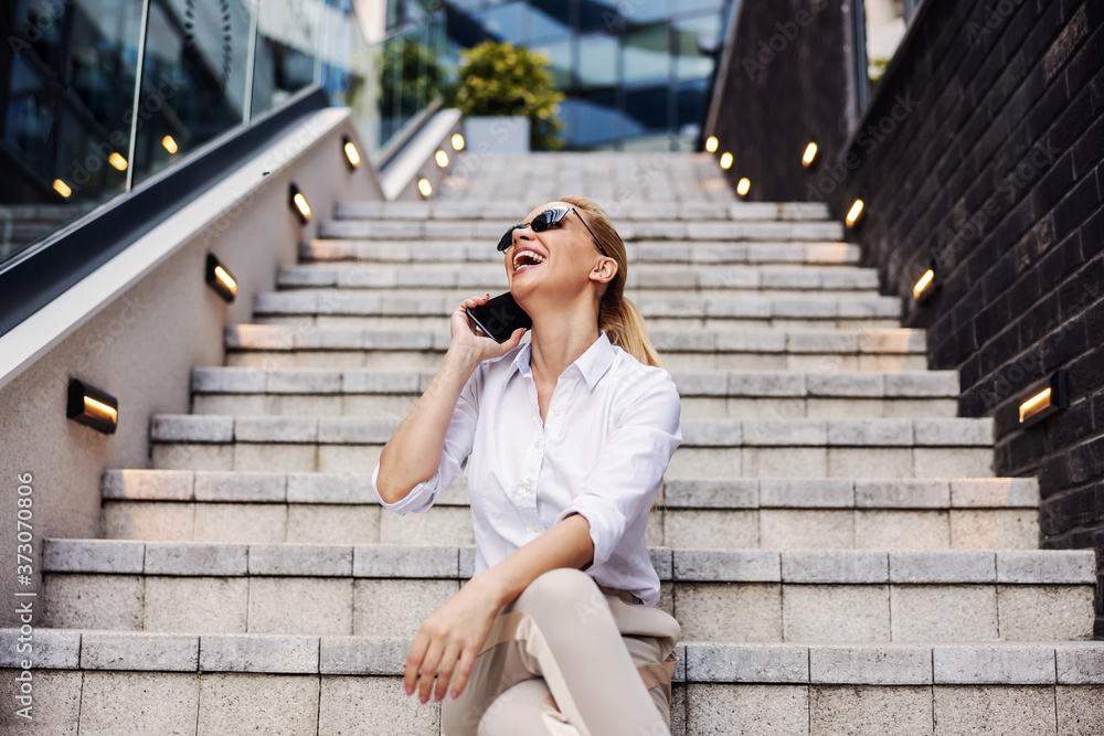 Smiling gorgeous blond fashionable businesswoman sitting on the stairs in front of business center and using smart phone.