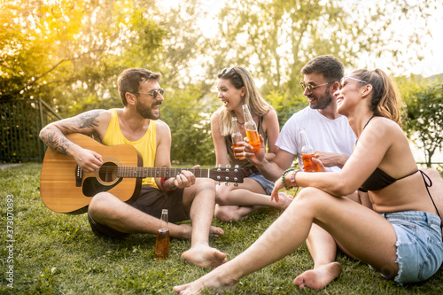 Two young couples sitting in the garden, enjoying and drinking beer on a sunny day while one of the men plays an acoustic guitar