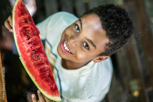 A bird's eye view image of a boy sitting on the bench, eating watermelon and looking at camera