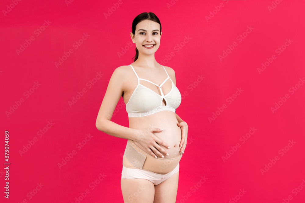 Portrait of pregnant woman in underwear wearing elastic bandage against pain in the back at pink background with copy space. Orthopedic abdominal support belt concept Portrait of pregnant woman in underwear wearing elastic bandage against pain in the back at pink background with copy space. Orthopedic abdominal support belt concept