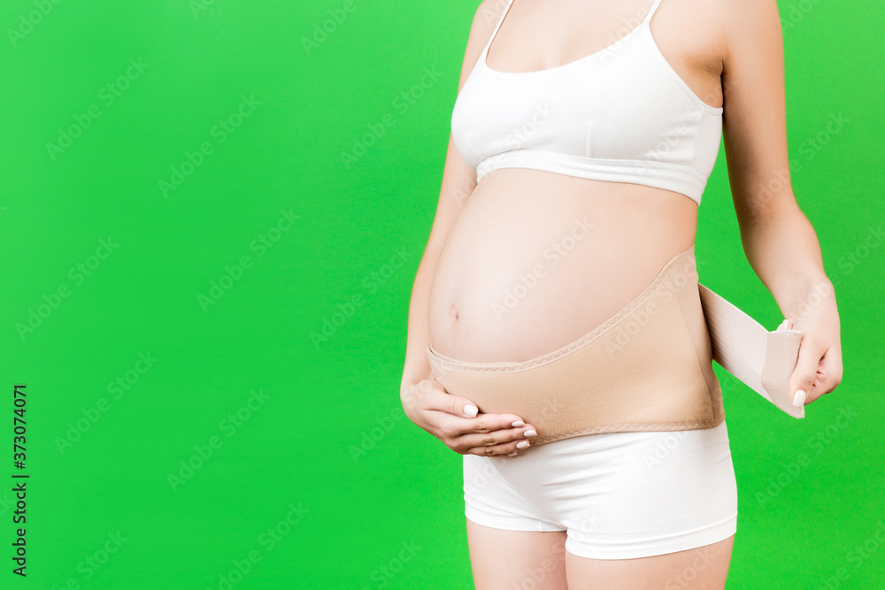 Close up of pregnant woman in underwear dressing maternity belt against pain in the back at green background with copy space. Orthopedic abdominal support belt concept Close up of pregnant woman in underwear dressing maternity belt against pain in the back at green background with copy space. Orthopedic abdominal support belt concept