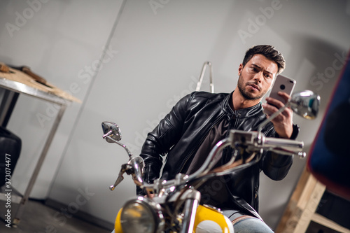 A beautiful muscular biker leads a video blog and talks on a video call on a smartphone while sitting on a motorcycle in the garage