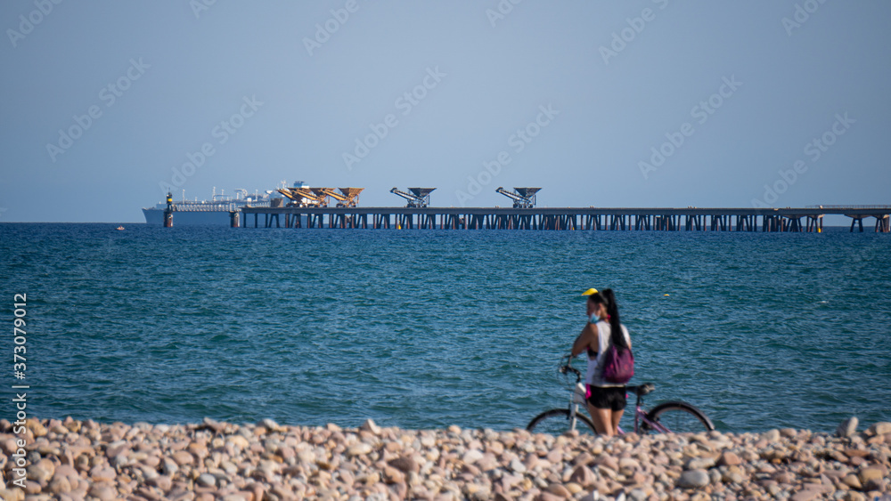 Mujer ciclista con mascarilla descansando en una playa con el puerto de fondo