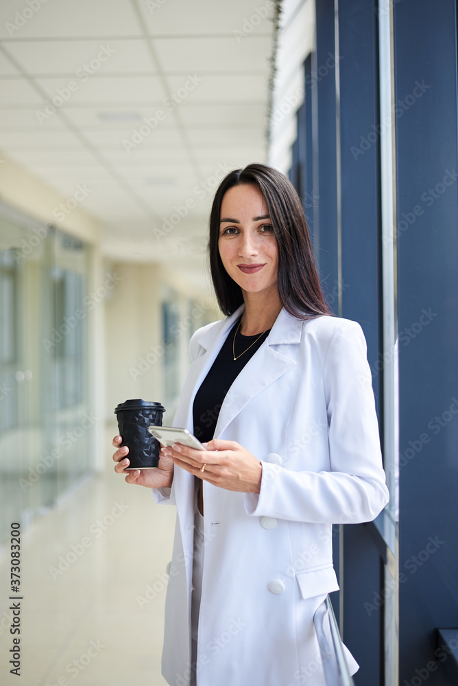Young brunette woman, wearing white business suit, holding cell phone and black paper coffee cup, standing in light office building near window. Businesswoman on lunch break. Corporate culture concept Young brunette woman, wearing white business suit, holding cell phone and black paper coffee cup, standing in light office building near window. Businesswoman on lunch break. Corporate culture concept