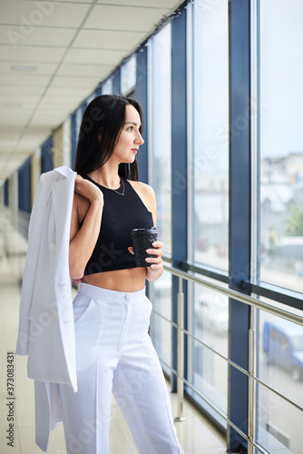 Young brunette woman, wearing white business with jacket in shoulder, holding black paper coffee cup, standing in light office building near window. Businesswoman on lunch break. Corporate culture. Free Download Young brunette woman, wearing white business with jacket in shoulder, holding black paper coffee cup, standing in light office building near window. Businesswoman on lunch break. Corporate culture. - GFXLoad