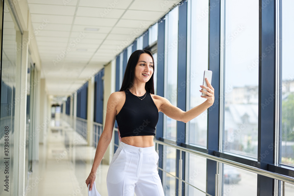 Young brunette woman, wearing white pants and black top, holding jacket in hand, taking selfie picture with cell phone in light passageway, posing for social media. Businesswoman on lunch break. Young brunette woman, wearing white pants and black top, holding jacket in hand, taking selfie picture with cell phone in light passageway, posing for social media. Businesswoman on lunch break.