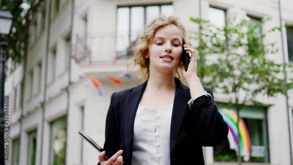 Walking down on the street and speaking on the phone businesswoman she have a curly blonde hair and holding digital tablet Walking down on the street and speaking on the phone businesswoman she have a curly blonde hair and holding digital tablet