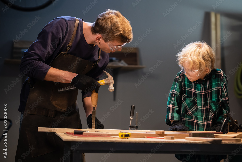 a young male carpenter is teaching woodwork to a young boy in his workshop. a young male carpenter is teaching woodwork to a young boy in his workshop.