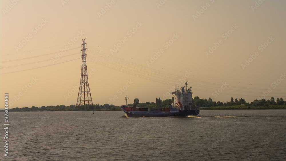 Elbe river near Hamburg harbor with huge power line and a ferry sailing by to deliver green energy from wind turbines during warm sunset, Germany