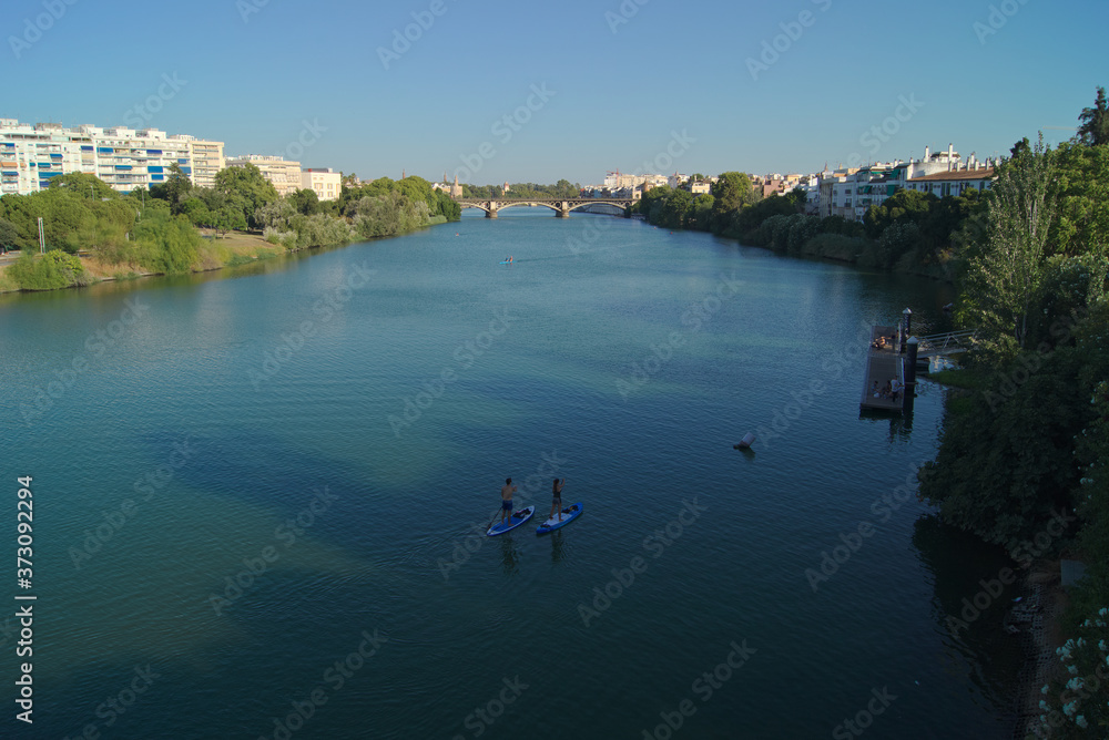 Vistas del río Guadalquivir a su paso por la ciudad de Sevilla con personas haciendo paddle surf y remo. Al fondo se puede ver el puente de Isabel II más conocido como puente de Triana.