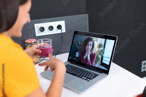 Beautiful young nutritionist woman having an online video call via laptop computer with a friend to showing detox handmade smoothie in the kitchen at home.