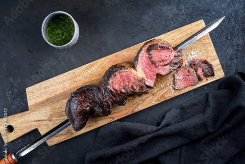 Barbecue dry aged wagyu Brazilian picanha from the sirloin cap of rump beef with chimichurri sauce sliced and offered as top view on a skewer on a wooden design board on rustic black background