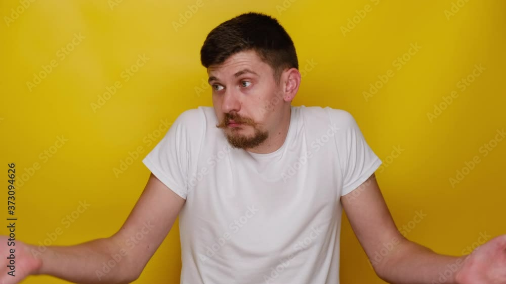 Indecisive young man keeps hands sideways, shrugs shoulders, guy cannot answer question, dressed in white t-shirt, has no idea, isolated on yellow studio background. people emotion concept Indecisive young man keeps hands sideways, shrugs shoulders, guy cannot answer question, dressed in white t-shirt, has no idea, isolated on yellow studio background. people emotion concept