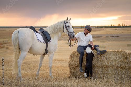 Man sitting on a straw bundle with his saddled horse and dog. Country scene. © fuen30